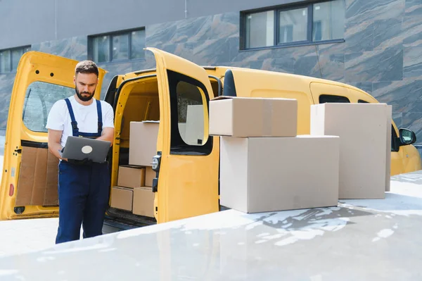 Kurye, paket dolu sarı minibüsünün yanındaki laptopta teslimat bilgilerini kontrol ediyor.