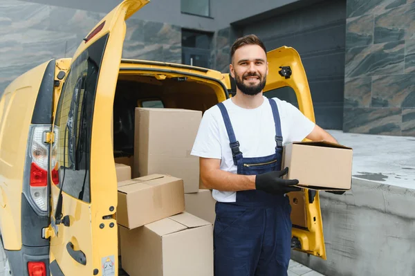 Eldivenli kurye, karton kutularla dolu sarı minibüsünün yanında bir paket tutuyor, gülümsüyor.