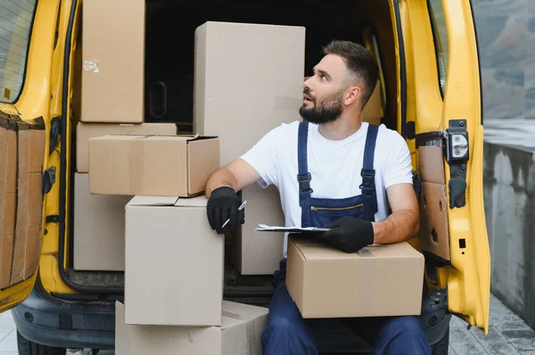Sarı minibüsünün arkasında oturan eldiven ve tulum giyen kurye karton kutulara bakıyor.