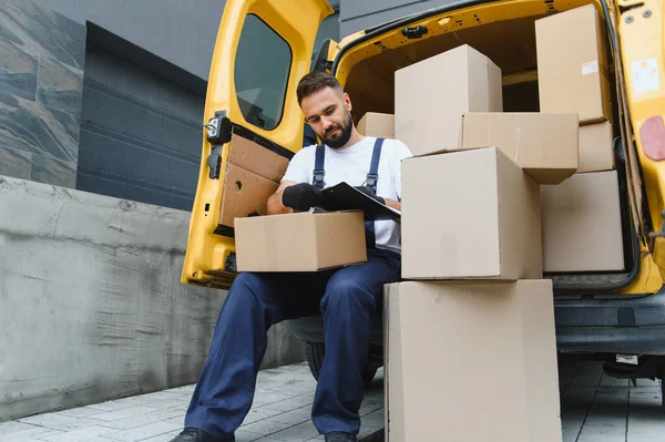 Eldiven giyen ve üniformalı bir kurye, sarı bir minibüste otururken karton kutuları kontrol ediyor.