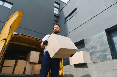 Kurye, açık teslimat minibüsünden karton kutu taşıyor. Paketi modern binanın önüne teslim ediyor.
