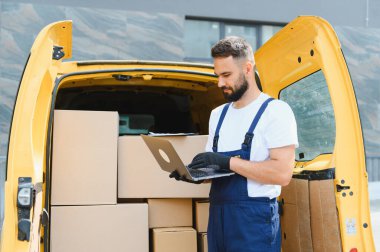Sevkiyat ve taşıma için karton kutularla dolu sarı bir minibüsün yanında dururken bir dizüstü bilgisayar kullanarak lojistiği yöneten teslimatçı.