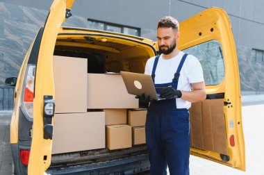 Kutularla dolu sarı bir minibüsün yanında dizüstü bilgisayarda çalışan teslimatçı. Lojistiği yönetiyor ve etkili paket dağıtımını sağlıyor.