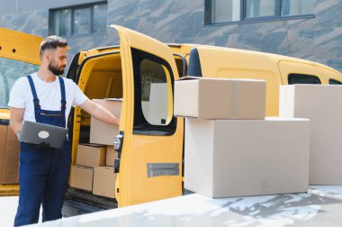 Tulum giyen kurye, gün ışığında parlak sarı bir minibüsü yüklerken dizüstü bilgisayardaki paketleri kontrol ediyor.