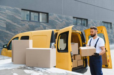 Eldiven takan kurye, modern bir binanın yanındaki sarı bir minibüse karton kutular yüklüyor.