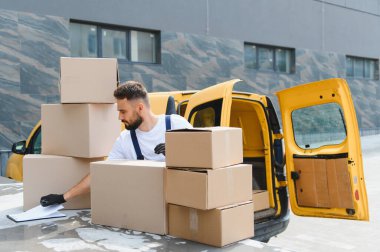 Tulum ve eldiven giyen kurye teslimat kamyonunun rampasındaki karton kutuları kontrol ediyor, elinde bir dosya tutuyor.