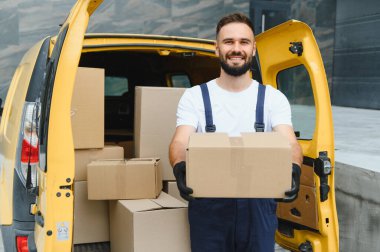Elinde karton bir kutu olan gülümseyen teslimatçı paketlerle dolu sarı bir minibüsün önünde duruyor.