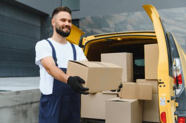 Eldiven takan gülümseyen teslimatçı, sarı kargo kamyonetinin yanında bir karton kutu taşıyordu.