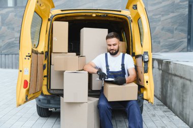 Eldiven takan kurye, karton kutularla dolu sarı minibüste oturuyor. Dosyada nakliye listesini kontrol ediyor.