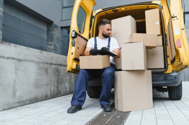 Eldiven takan teslimatçı, karton kutularla dolu bir minibüste oturuyor. Verimli sipariş yerine getirilmesi için paket listesini kontrol ediyor.