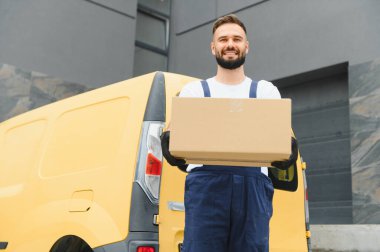 Eldiven takan gülümseyen teslimatçı, sarı minibüsünün yanında bir karton kutu tutuyor, nakliyeye hazır.