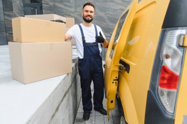 Tulumlu ve eldivenli bir kurye. Başparmak kaldırıyor. Karton kutuların yanında duruyor.