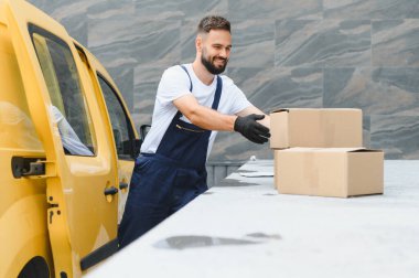 Gülümseyen teslimatçı, parlak sarı bir minibüsten karton kutuları indiriyor, eldiven ve tulum giyiyor, profesyonel bir hizmet sergiliyor.