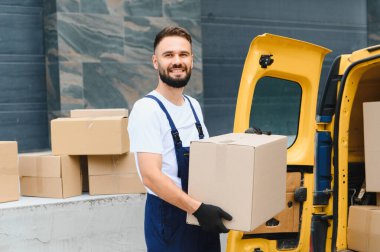 Tulum ve eldiven giymiş kurye, açık bir teslimat minibüsünün yanında bir karton kutu tutarken gülümsüyor, bir sonraki sevkiyat için hazır.
