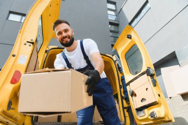 Eldiven giyen kurye, açık sarı bir minibüsün yanında güpegündüz dururken karton bir kutuyu etkili bir şekilde tutuyor.