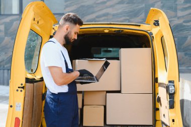 Tulum ve eldiven giyen kurye, dizüstü bilgisayar kullanarak sarı bir minibüste karton kutuları kontrol ediyor.