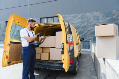 Eldiven takan kurye, bir dizüstü bilgisayarda çalışırken şehir caddesinde sarı bir minibüsün içindeki karton kutuları kontrol ediyor.