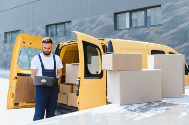 Kurye, karton kutularla dolu sarı minibüsünün yanındaki dizüstü bilgisayardan siparişleri kontrol ediyor.