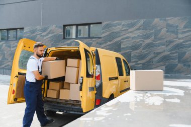 Karton kutuları parlak sarı bir minibüsten boşaltan teslimatçı, kentsel bir ortamda dinamik nakliye ve lojistik süreci vurguluyor.