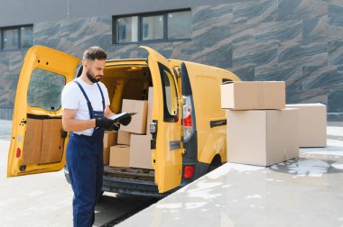 Eldiven takan kurye, karton kutularla dolu sarı bir minibüsün yanındaki paketleri kontrol ediyor, sevkiyat ve paket yönetiyor.