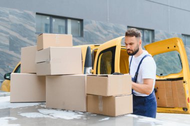 Eldiven takan kurye, yoğun bir iş gününde parlak sarı bir teslimat kamyonunun çatısına istiflenmiş karton kutuları inceliyor.