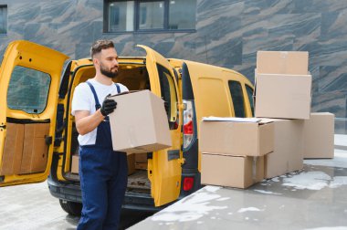Eldiven giyen kurye, açık teslimat kamyonundan karton kutu taşıyor. Yerde daha fazla paket var.