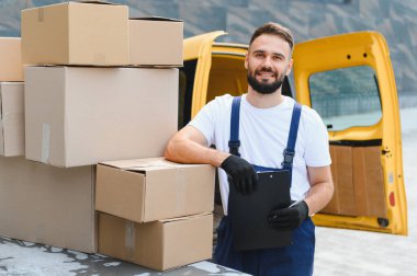 Kurye teslimat listesini panoda, açık teslimat kamyonetinin yanındaki karton kutuların yanında kontrol ediyor.