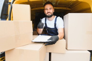 Minibüsün içinde karton kutular, hızlı teslimat ve paket taşıma konsepti ile çevrili bir şekilde teslimat notunu tutarken kurye başparmaklarını gösteriyor