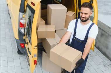 Gülümseyen teslimatçı, sarı bir minibüsten eldiven ve tulum giymiş, karton kutuları boşaltıyor.