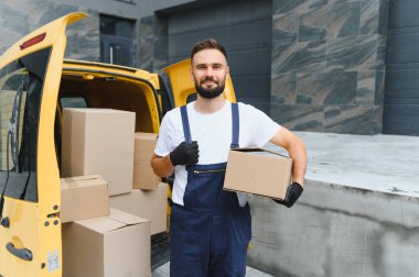 Karton kutularla dolu bir kargo aracının yanında gururla duran bir paket taşıyan ve başparmak kaldıran kurye, başarıya doğru yol alıyor.