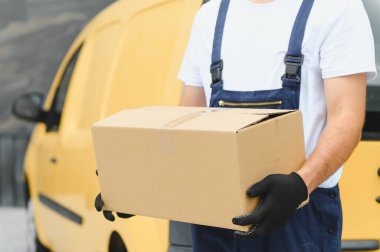 Sarı minibüsün yanında karton kutu taşıyan tulum ve eldiven giyen kurye, paket teslimat servisi konsepti.