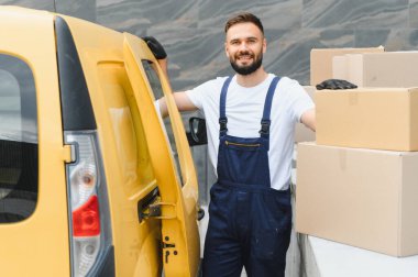 Kurye tulum ve eldiven giymiş, karton kutuların üzerine yaslanmış, teslimat minibüsünün yanına.