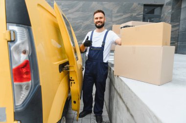 Üniformalı ve eldivenli bir kurye, sarı teslimat aracının yanında teslim edilmeye hazır paketler ve kapılar ile baş parmak işareti yapıyor.
