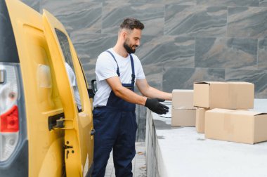Eldiven takan kurye, sarı bir kargo aracının yakınındaki karton kutuları kontrol ediyor. Malların nakliyesi ve taşınması için hazırlanıyor.