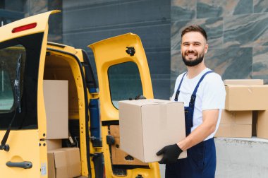 Kurye tulumlu ve siyah eldivenli sarı bir minibüsün yanında bir karton kutu tutuyor, gülümsüyor.