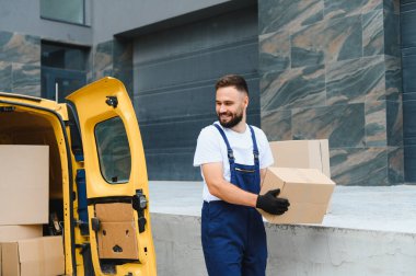 Sarı minibüsten karton kutu taşıyan gülümseyen teslimatçı, şehre paketler getiriyor.