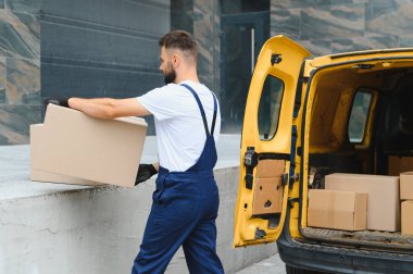 Kurye, açık bir teslimat minibüsünden karton bir kutu taşıyor, müşterilere paket taşırken şehir sokaklarında geziniyor.