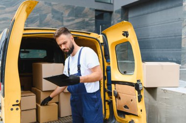 Eldiven ve tulum giyen kurye karton kutuları kontrol ederken teslimat minibüsünde pano tutuyor.