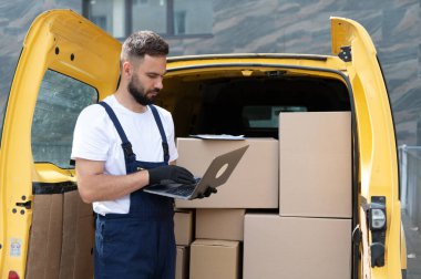 Karton kutularla dolu, verimli nakliye için hazır sarı bir minibüsün yanında duran bir dizüstü bilgisayardaki siparişleri kontrol eden kurye.