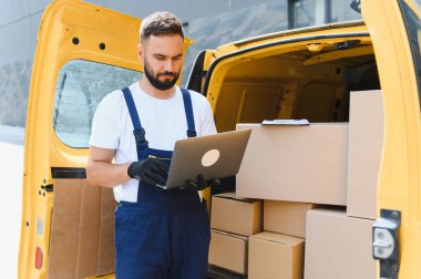 Karton kutularla dolu sarı bir minibüsün yanına park edilmiş bir dizüstü bilgisayardaki siparişleri kontrol eden kurye.