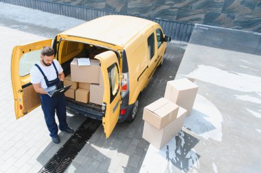 Kurye, karton kutularla dolu sarı minibüsünün yanındaki laptopta teslimat bilgilerini kontrol ediyor.