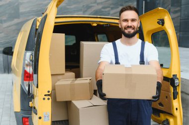 Paketlerle dolu sarı bir minibüsün yanında duran karton bir kutu taşıyan gülümseyen teslimatçı.