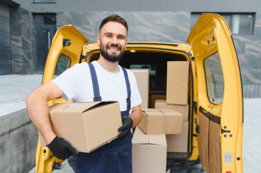 Eldiven takan ve gülümseyen bir teslimatçı içi kutu dolu sarı bir minibüsün önünde nakliyeye hazır bir paket tutarken.