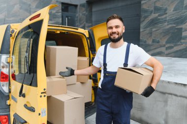 Sarı bir minibüste karton kutular sunan gülümseyen teslimatçı verimli ve güvenilir bir nakliye hizmeti sunuyor.