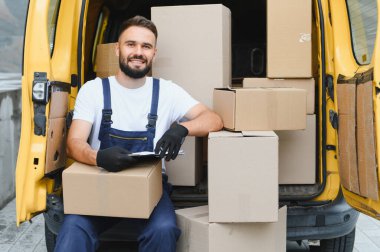 Karton kutulara bakan kurye, sarı bir minibüste elinde bir dosya ve eldiven tutarken...