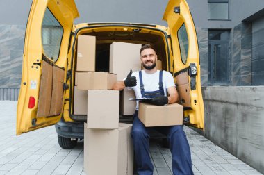 Kurye açık teslimat minibüsünde oturuyor, gülümsüyor ve karton kutularla çevrili başparmaklarını kaldırıyor.