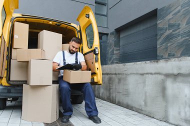 Eldiven giyen kurye teslimat listesini kontrol ediyor. Karton kutularla dolu açık bir minibüste oturuyor. Binanın önündeki sevkiyatı yönetiyor.