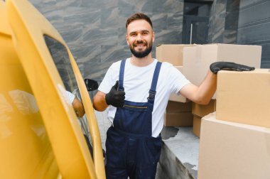 Tulum ve eldiven giyen kurye, karton kutular ve sarı teslimat minibüsünün yanında baş parmak işareti yapıyor.