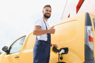 Kurye, modern bir binanın yakınında elektrik minibüsünü şarj ederken, sürdürülebilir taşımacılığa olan bağlılığı vurguluyor