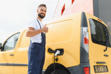 Elektrikli minibüsü şarj ederken başparmaklarını gösteren kurye, sürdürülebilir dağıtım uygulamalarını kucaklıyor